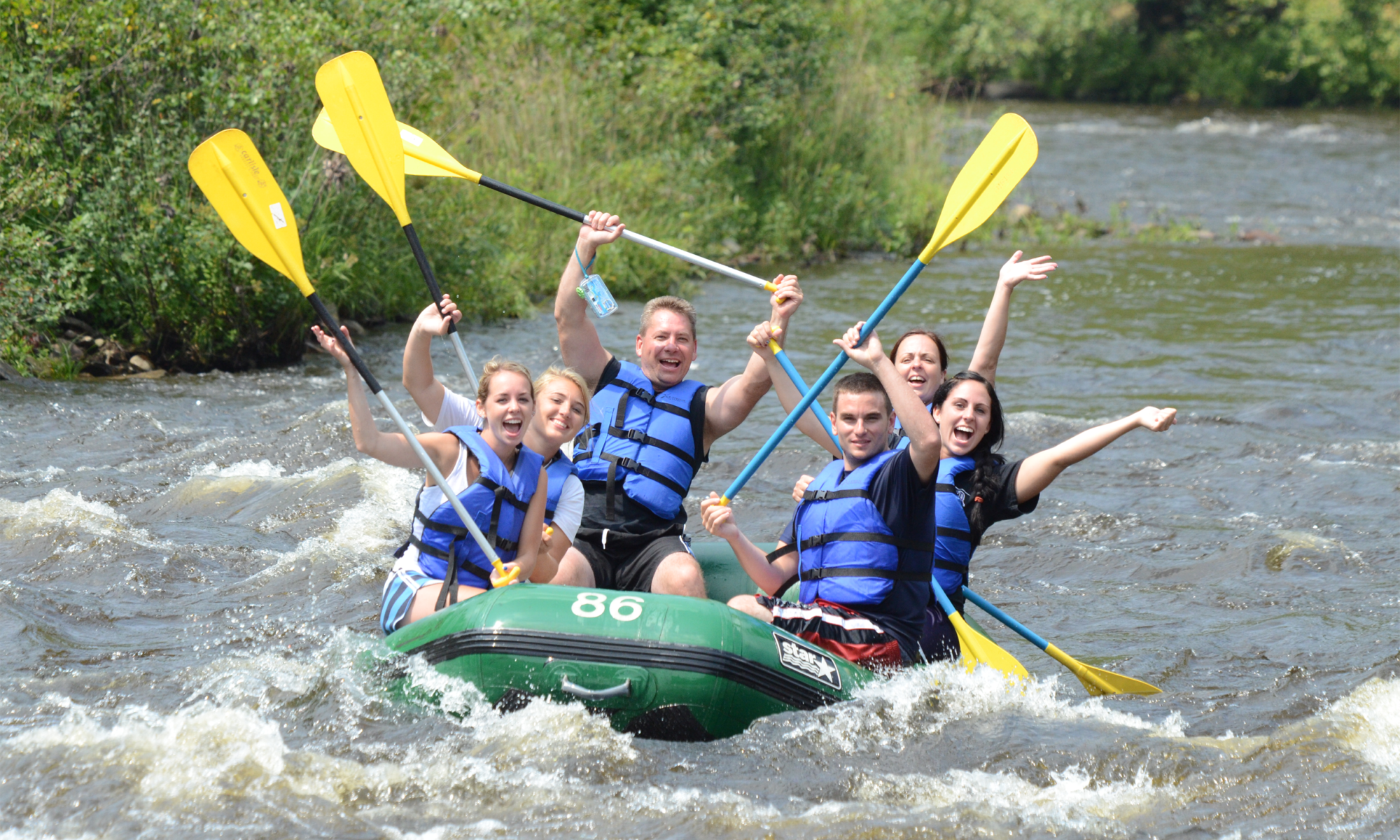 Group of six people in life vests cheerfully rafting on a river with yellow paddles.