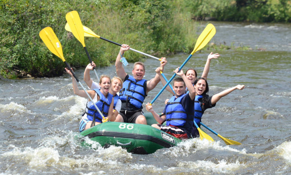Group of six people in life vests cheerfully rafting on a river with yellow paddles.