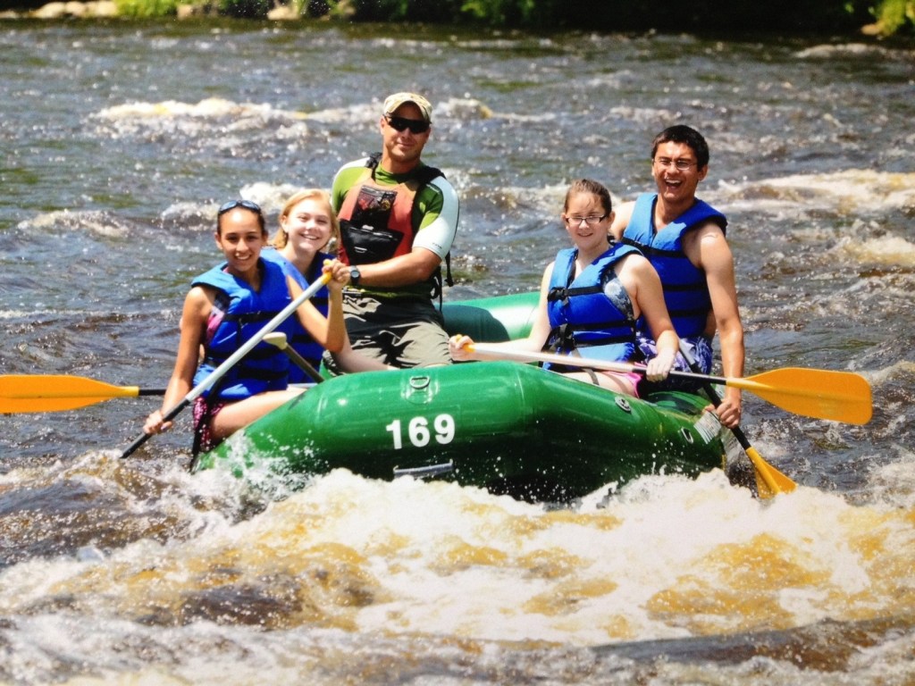 Group of people rafting on a river, wearing blue life jackets in a green raft.