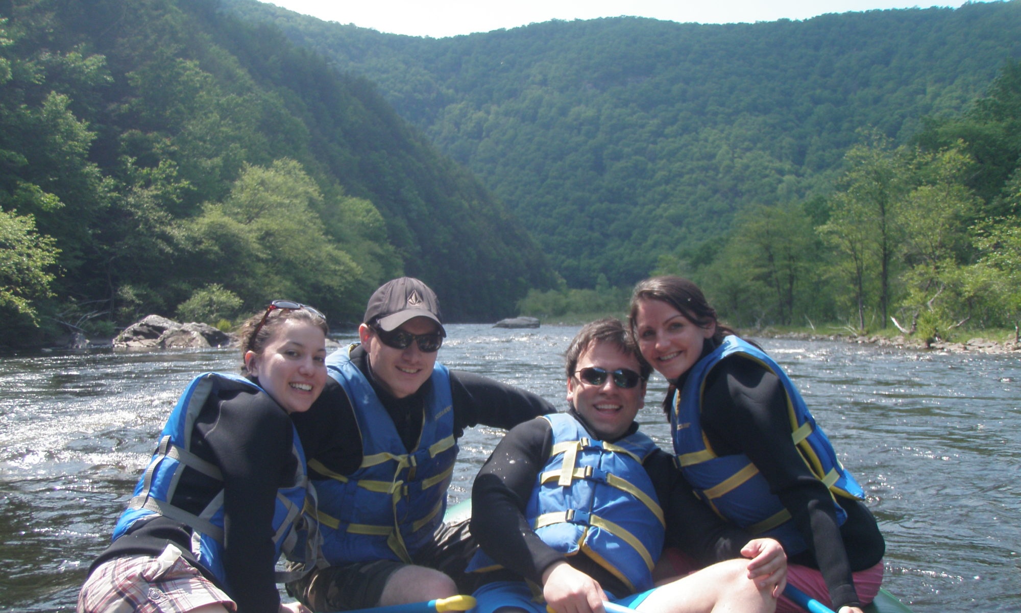 Four people rafting on a river surrounded by forested hills.
