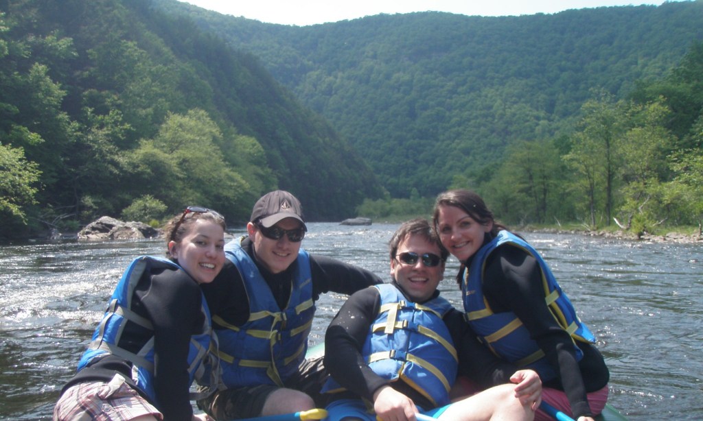 Four people rafting on a river surrounded by forested hills.