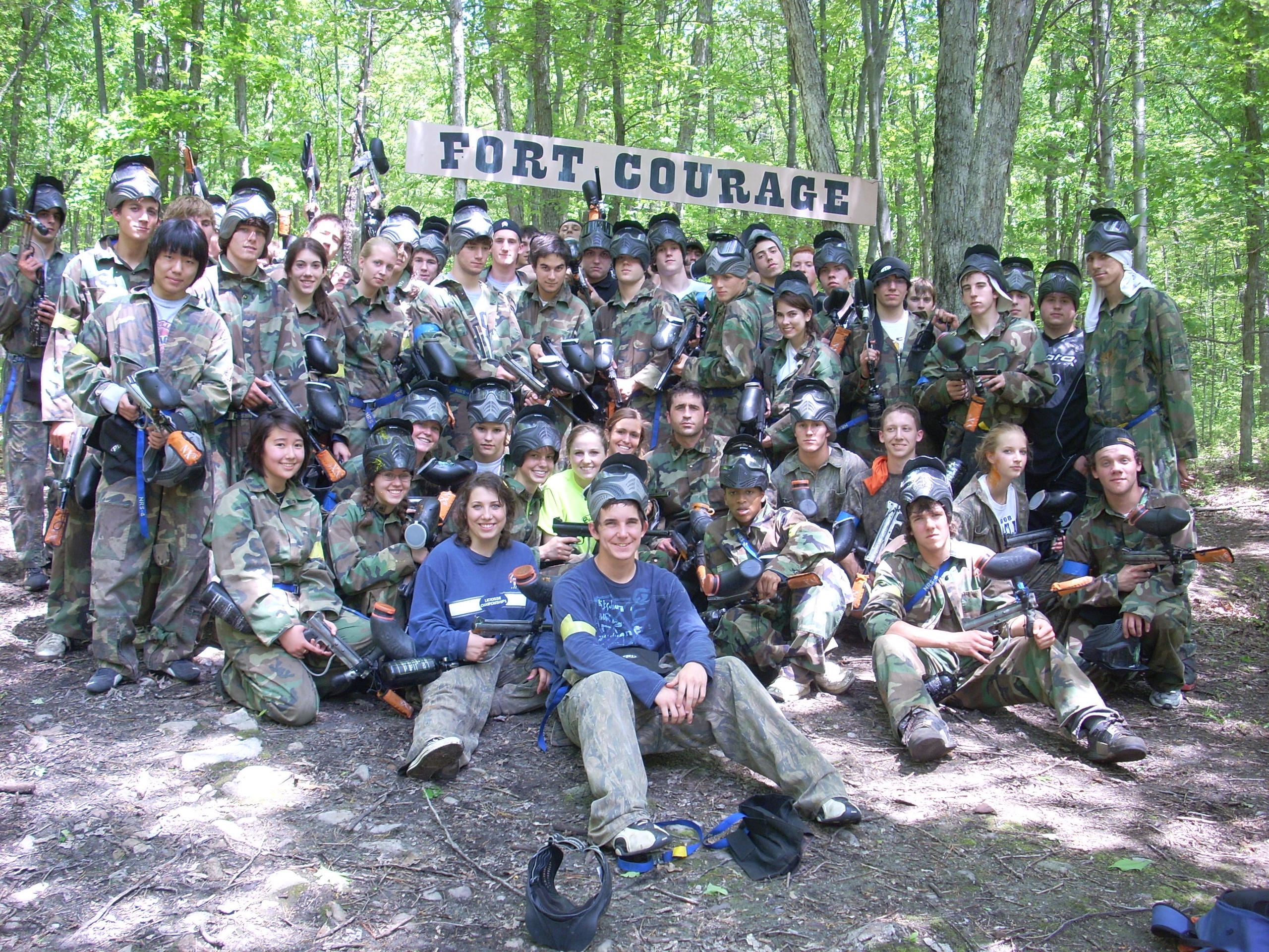 Group of people in camouflage holding paintball gear at Fort Courage, surrounded by trees.