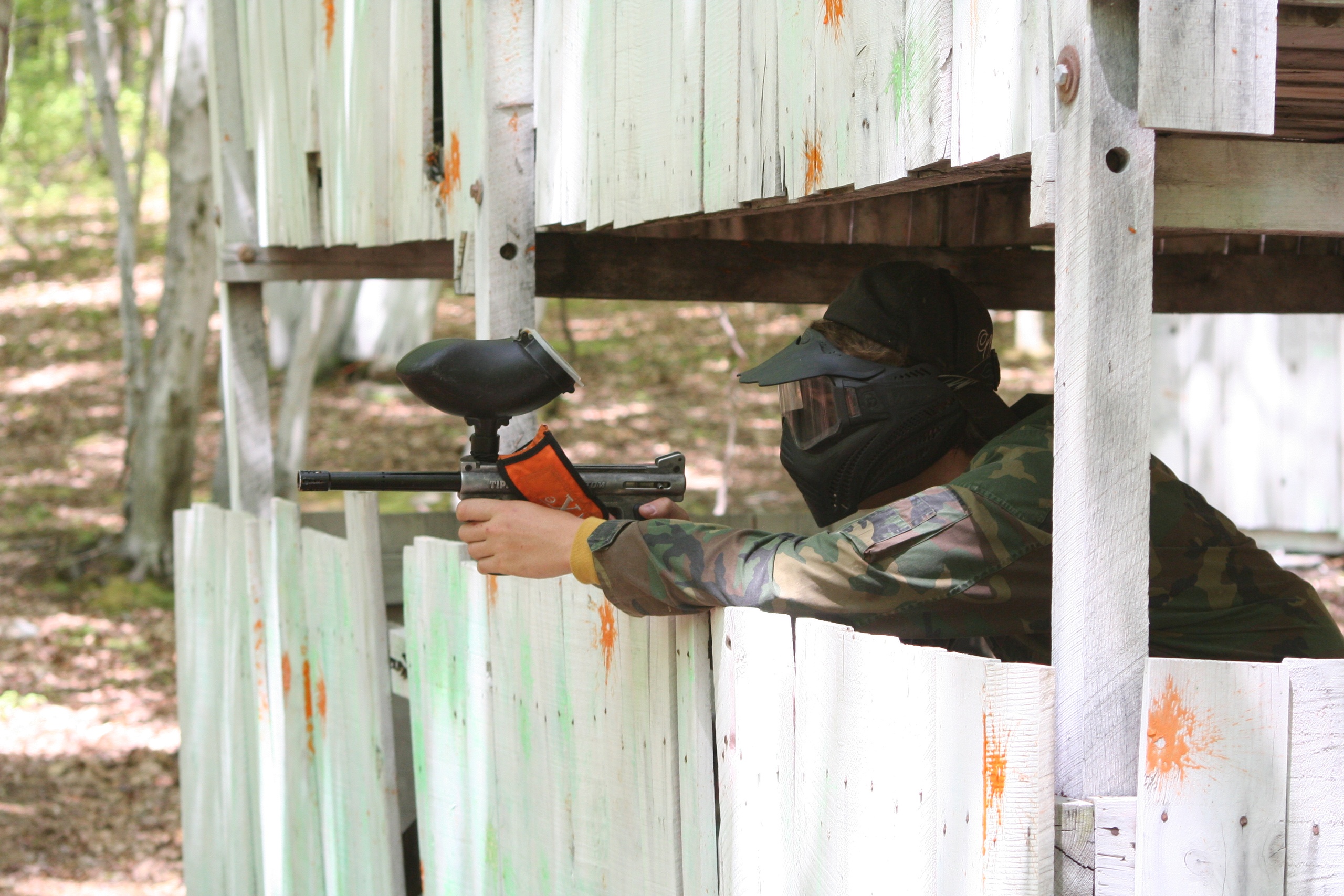 Person in camouflage with paintball gun at wooden barricade.