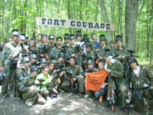 Group of people in camouflage gear holding paintball guns in forest under 'Fort Courage' sign.