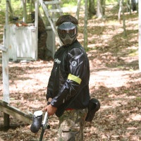 Person in paintball gear and mask holding marker in wooded area.