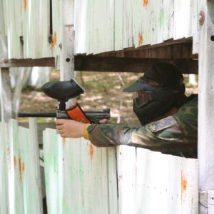 Person in camouflage playing paintball, aiming a marker through wooden slats.