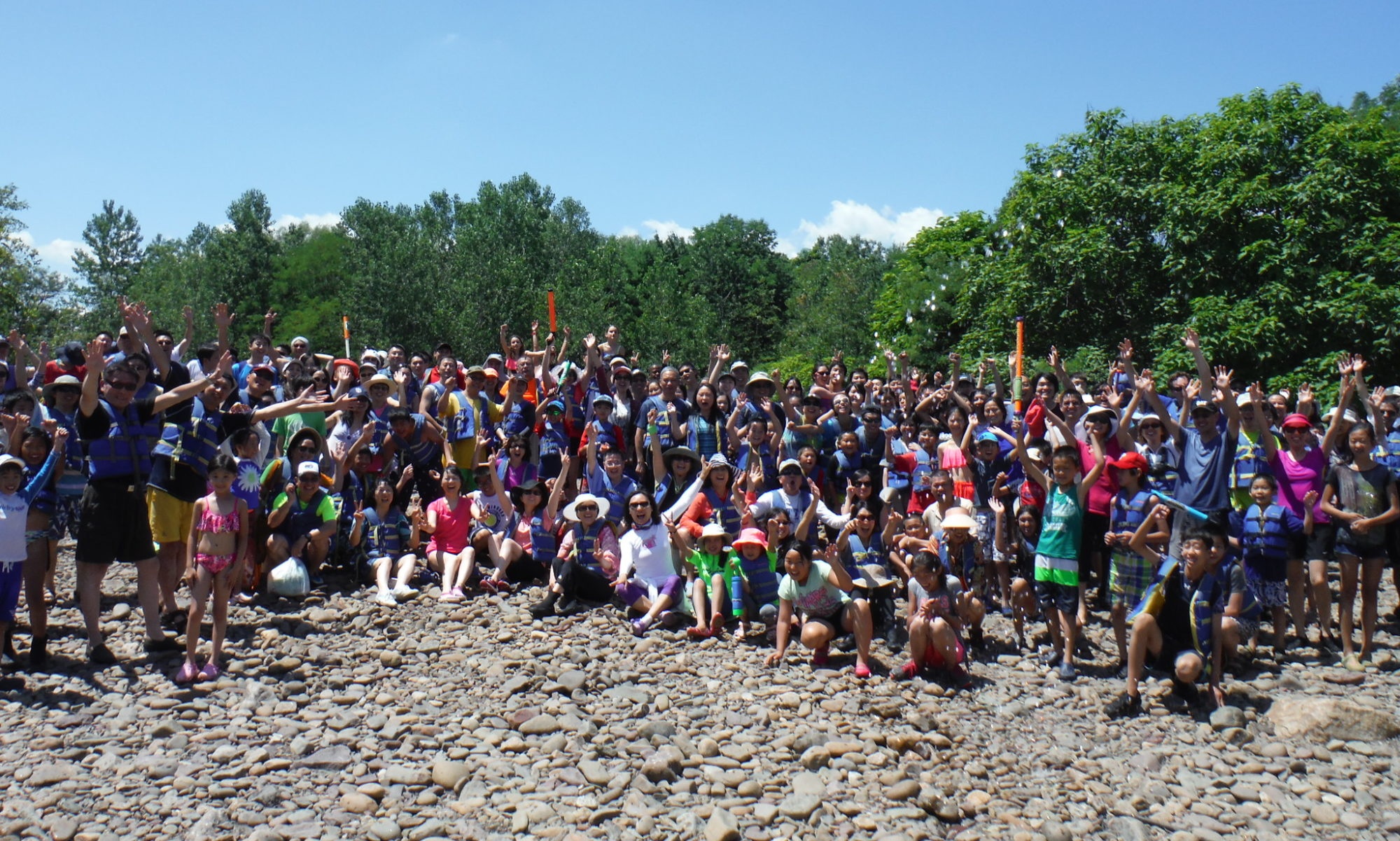 Large group of people on a rocky area, cheering with hands raised, trees in the background.