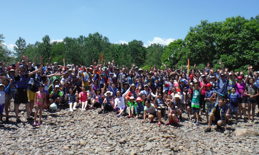 Large group of people on a rocky area, cheering with hands raised, trees in the background.
