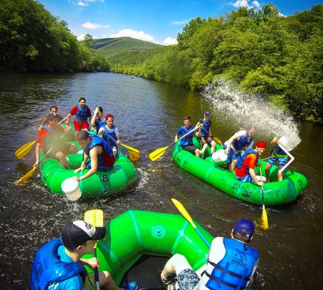 People rafting on river in green inflatable boats, splashing water, surrounded by green trees.