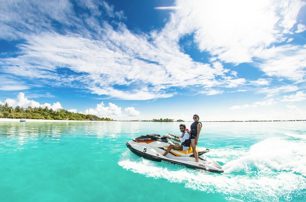 Two people riding a jet ski on clear turquoise water near a tropical beach.