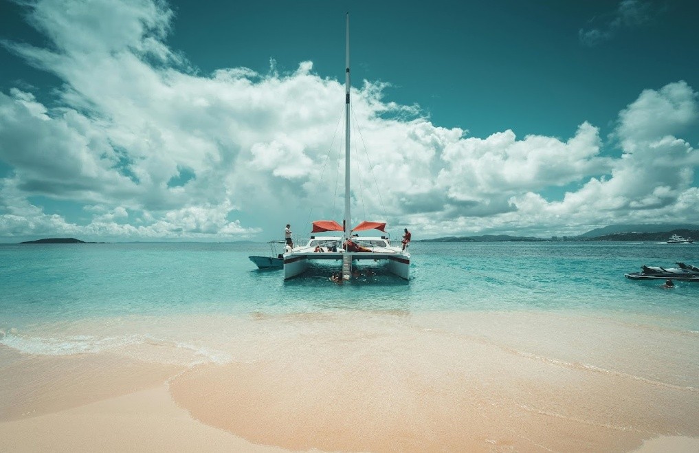 Catamaran moored on clear blue water near sandy beach under cloudy sky.