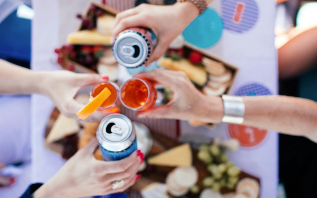 Four people toasting with drinks above a table with snacks.