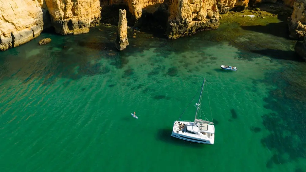 Catamaran and kayaker in clear water near rocky cliffs.