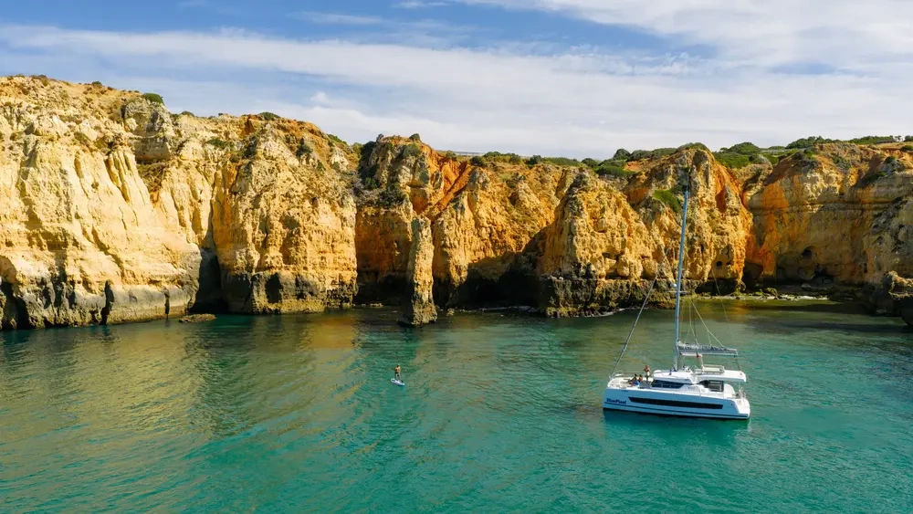 Sailboat near tall, rocky cliffs over clear blue-green water under a partly cloudy sky.
