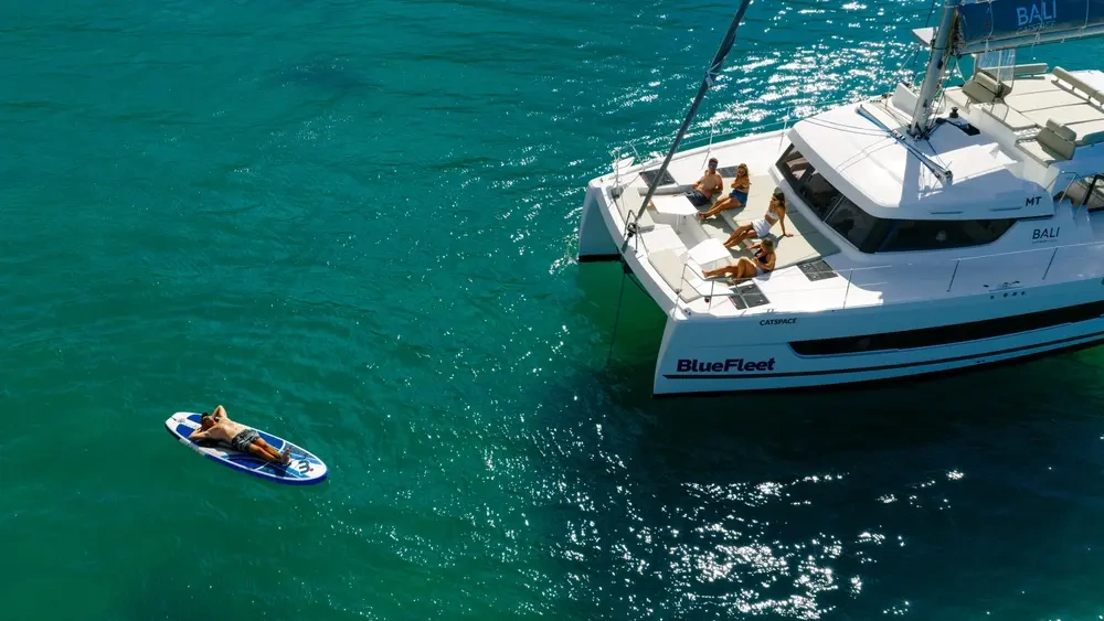 Catamaran and person on paddleboard in clear turquoise water.