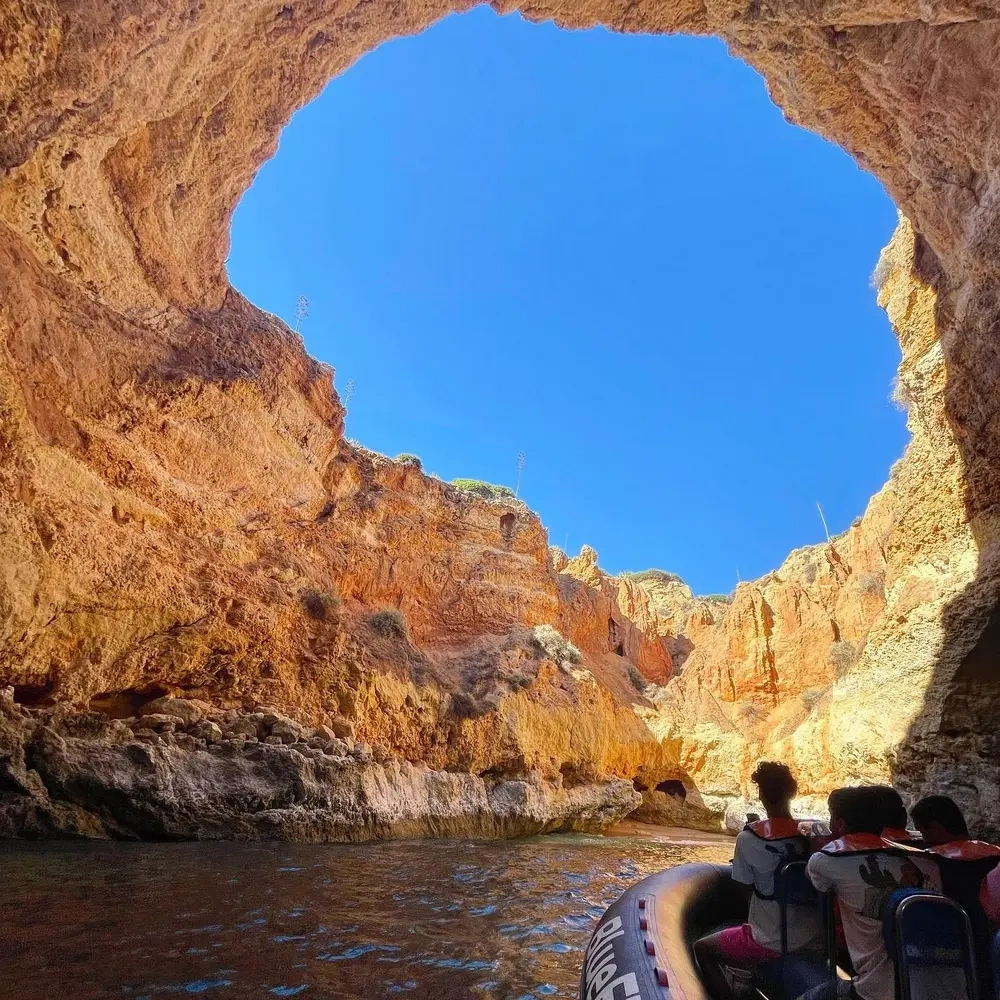 People in a boat inside a rocky cave with a large opening to the blue sky above.