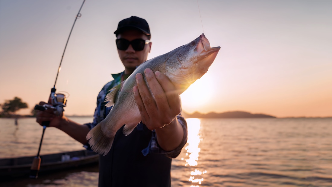 Person holding a fish in one hand with a fishing rod in the other, standing by a lake during sunset.