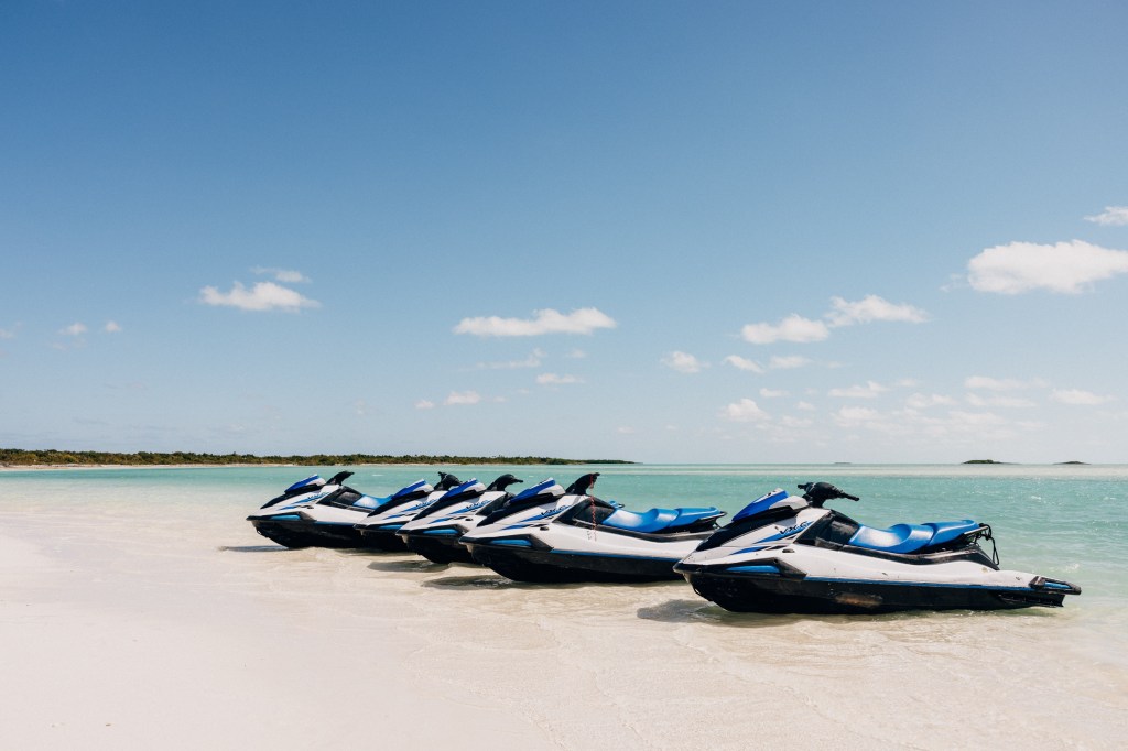 Five jet skis lined up on a sandy beach with clear blue water.