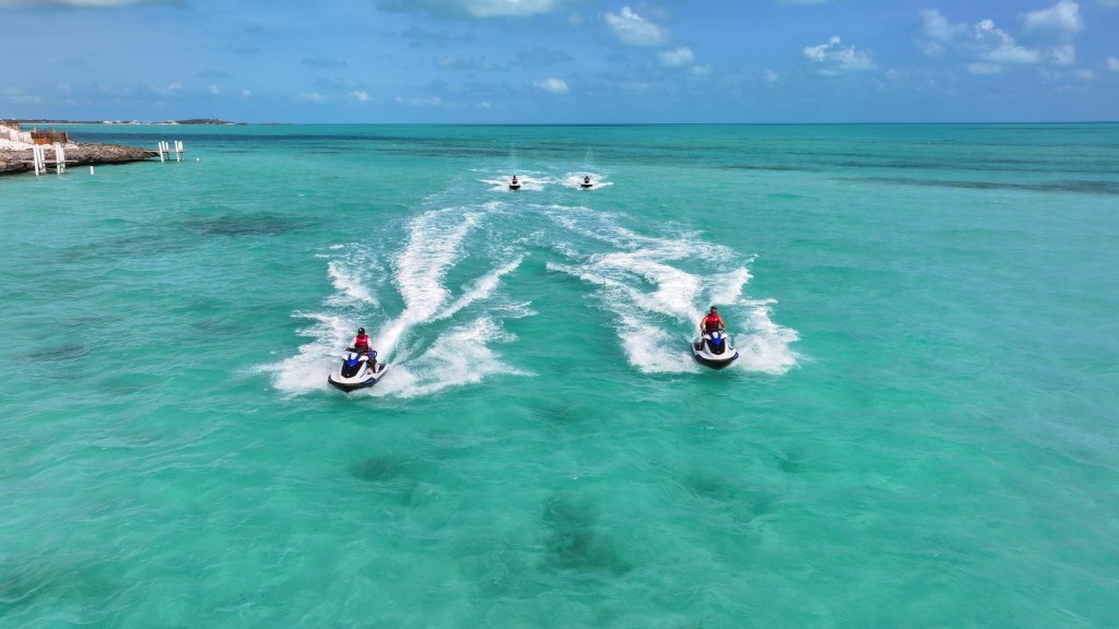 Four people riding jet skis on clear turquoise water under a blue sky.
