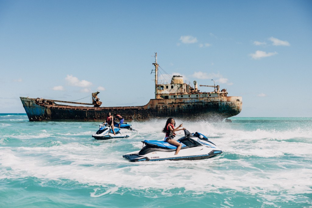 People riding jet skis near a large, rusty shipwreck in the ocean under a blue sky.
