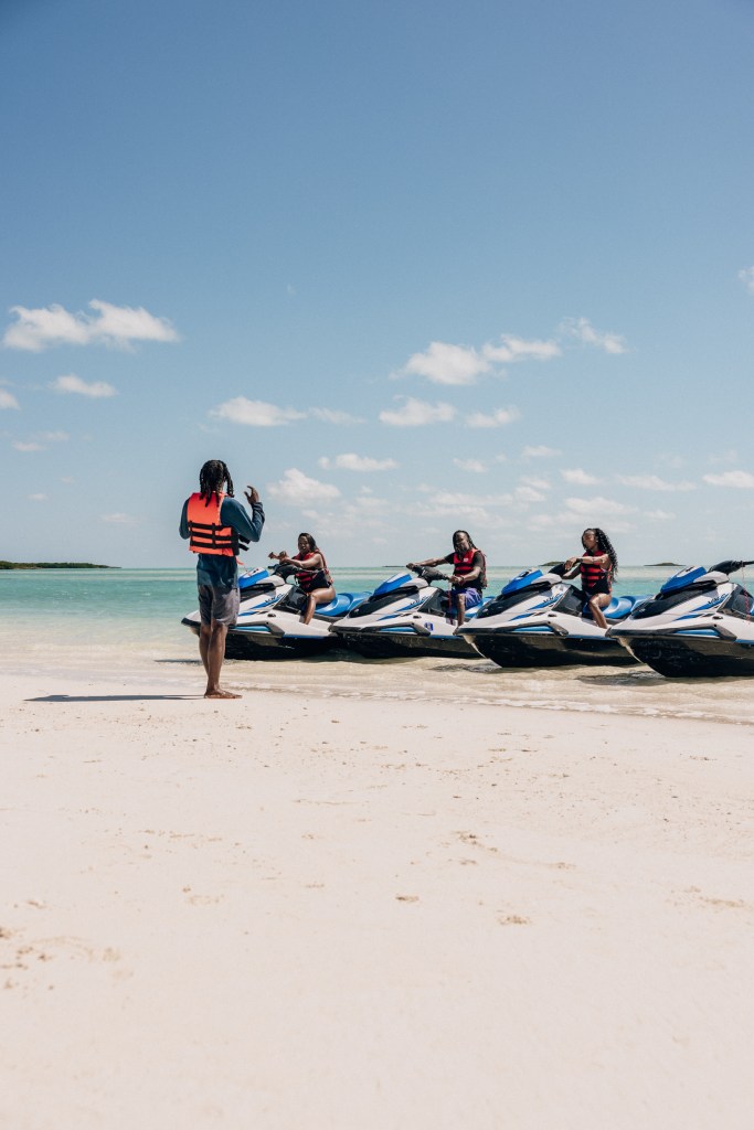 People on jet skis by a beach, one standing, under a clear sky.