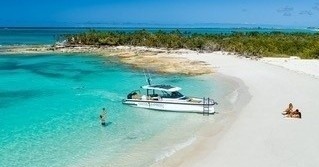 Boat by sandy beach, clear water, one person swimming, couple relaxing on shore, lush island in background.