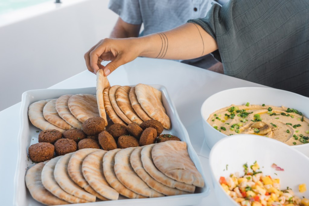 Person picking pita bread from tray with falafel, near bowls of hummus and salad.