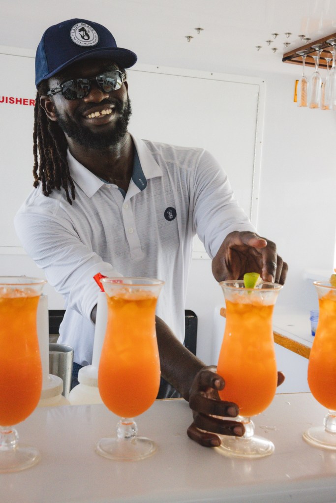 Man in hat serving orange cocktails in tall glasses, smiling on a boat.