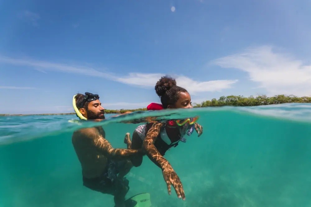 Man assisting woman snorkeling in clear blue water, split view above and below surface.