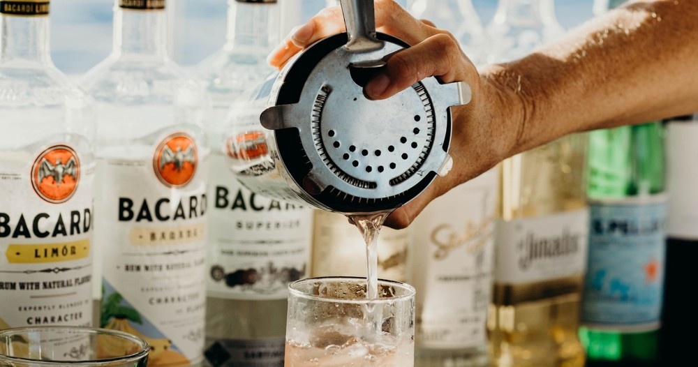 Hand pouring a drink from shaker into glass, Bacardi bottles in background.