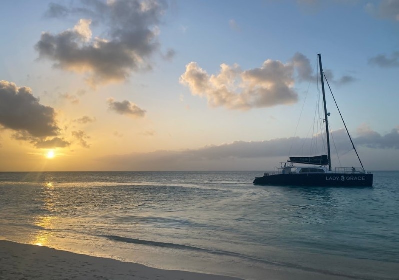 Sailboat on ocean at sunset with clouds, near sandy beach.