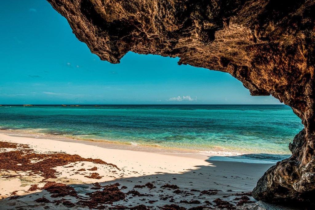 View of a sandy beach with turquoise waters, seen from under a rocky overhang.