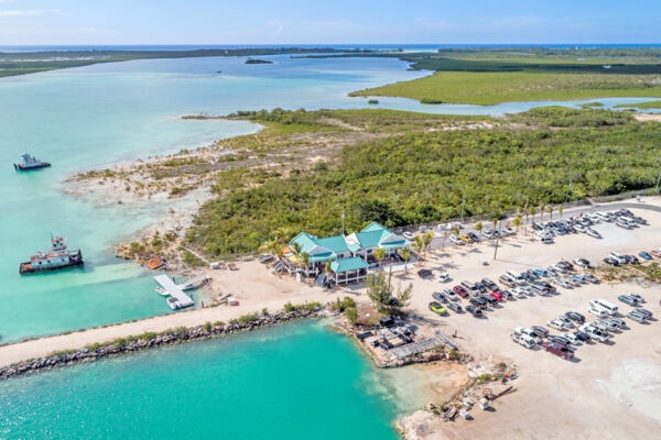 Aerial view of a coastal area with parked cars, a building, and two boats near turquoise water.