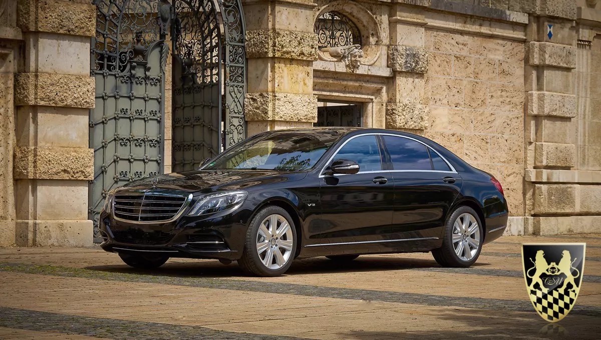 Black luxury sedan parked near ornate stone gate, emblem on wall.