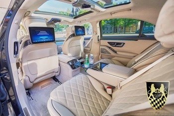 Luxurious car interior with screens, quilted seats, and sunroof, parked near trees and building.