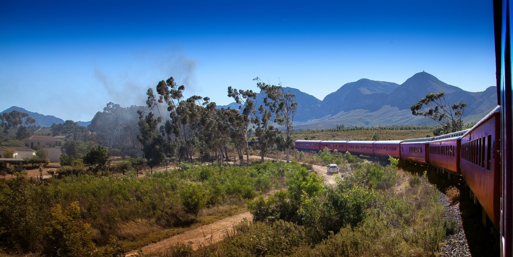 Train traveling through rural landscape with mountains in background.