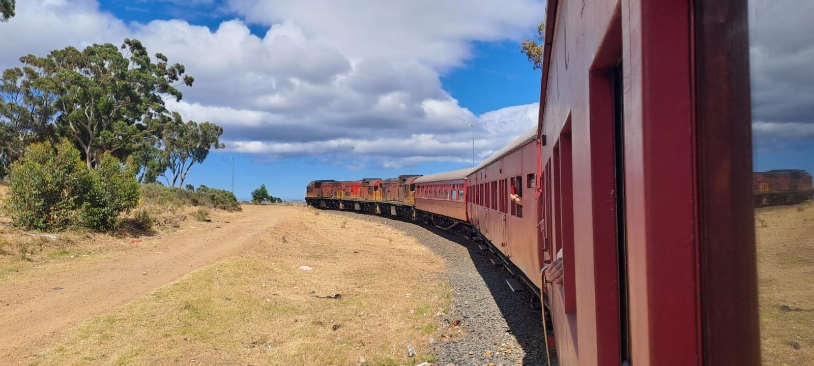 Red train on curved tracks in a rural landscape with trees and blue sky.