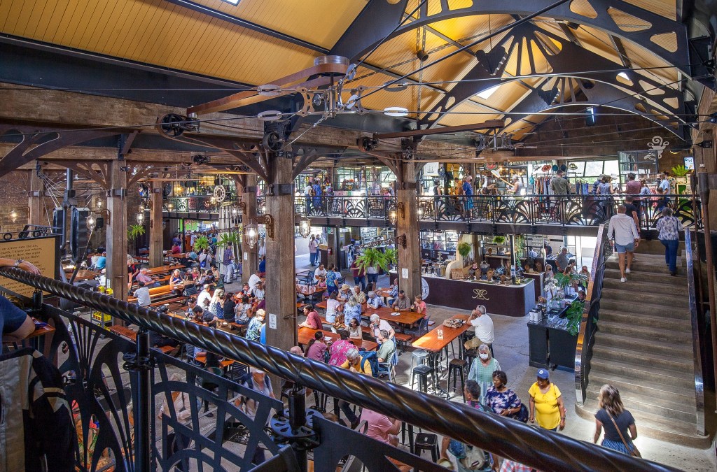 Busy indoor market with people, tables, iron beams, and wooden accents.