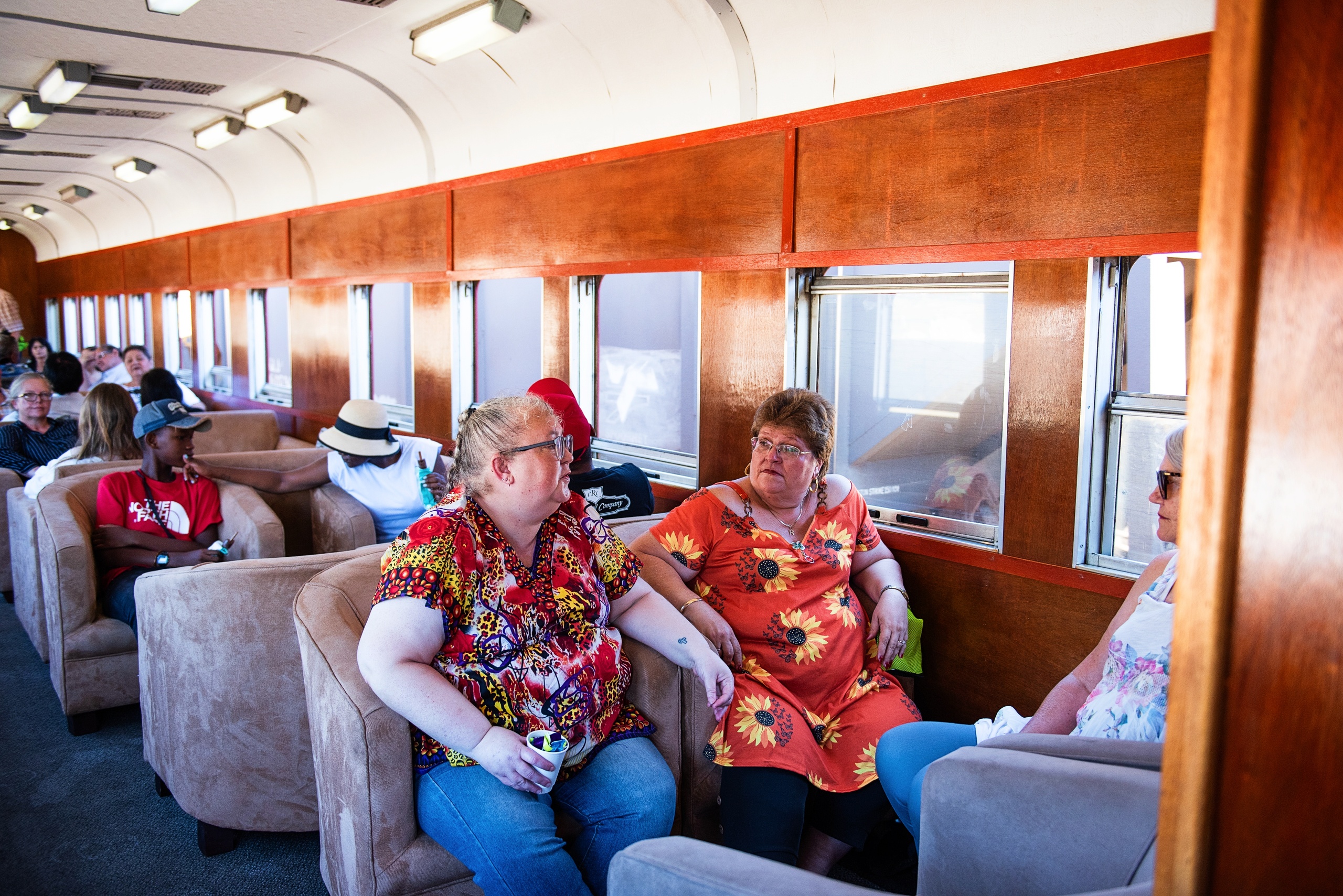 People sitting and talking inside a train with cushioned seats and wooden paneling.