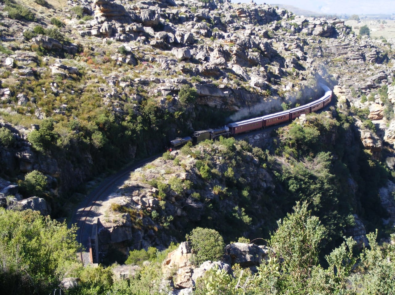 Train winding through rocky, hilly landscape with vegetation.