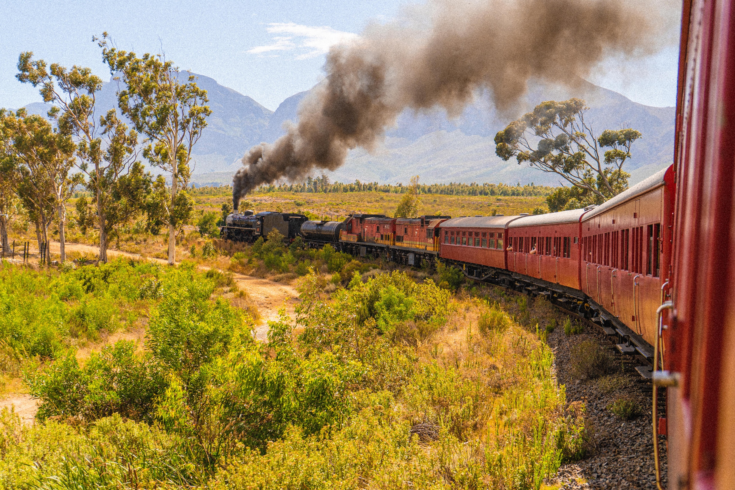 Steam train with red carriages traveling through countryside with mountains.