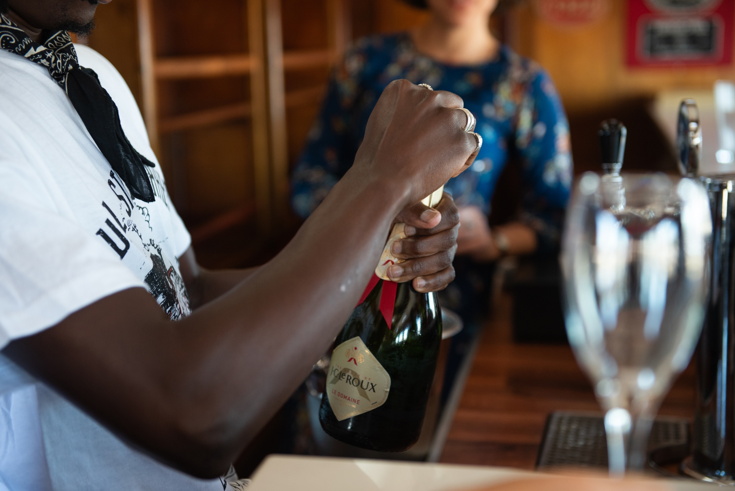 Person opening a champagne bottle near a bar counter with a wine glass nearby.