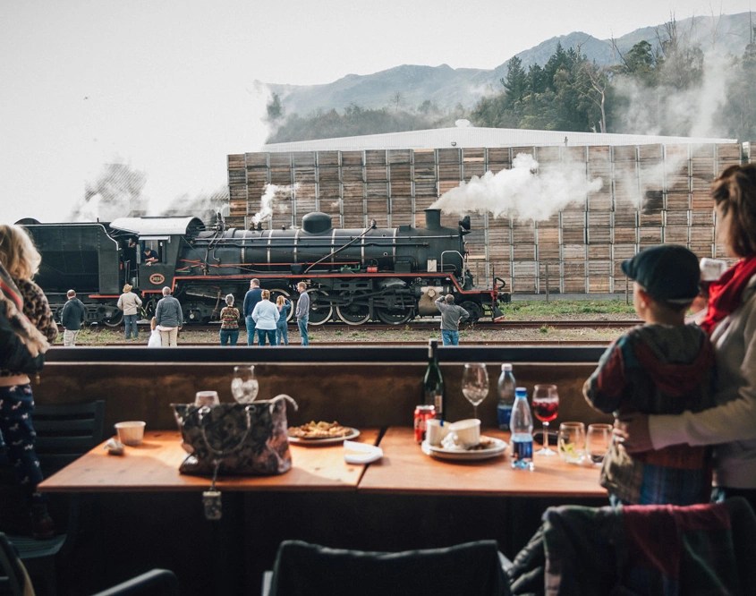 People watching a steam train from a restaurant with food and drinks on the table.