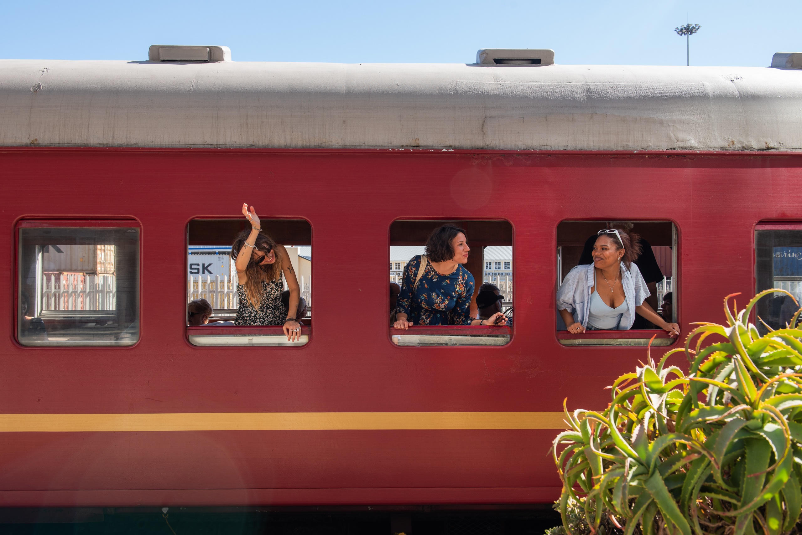 Three people leaning out of train windows, one is waving, in a stationary red train.