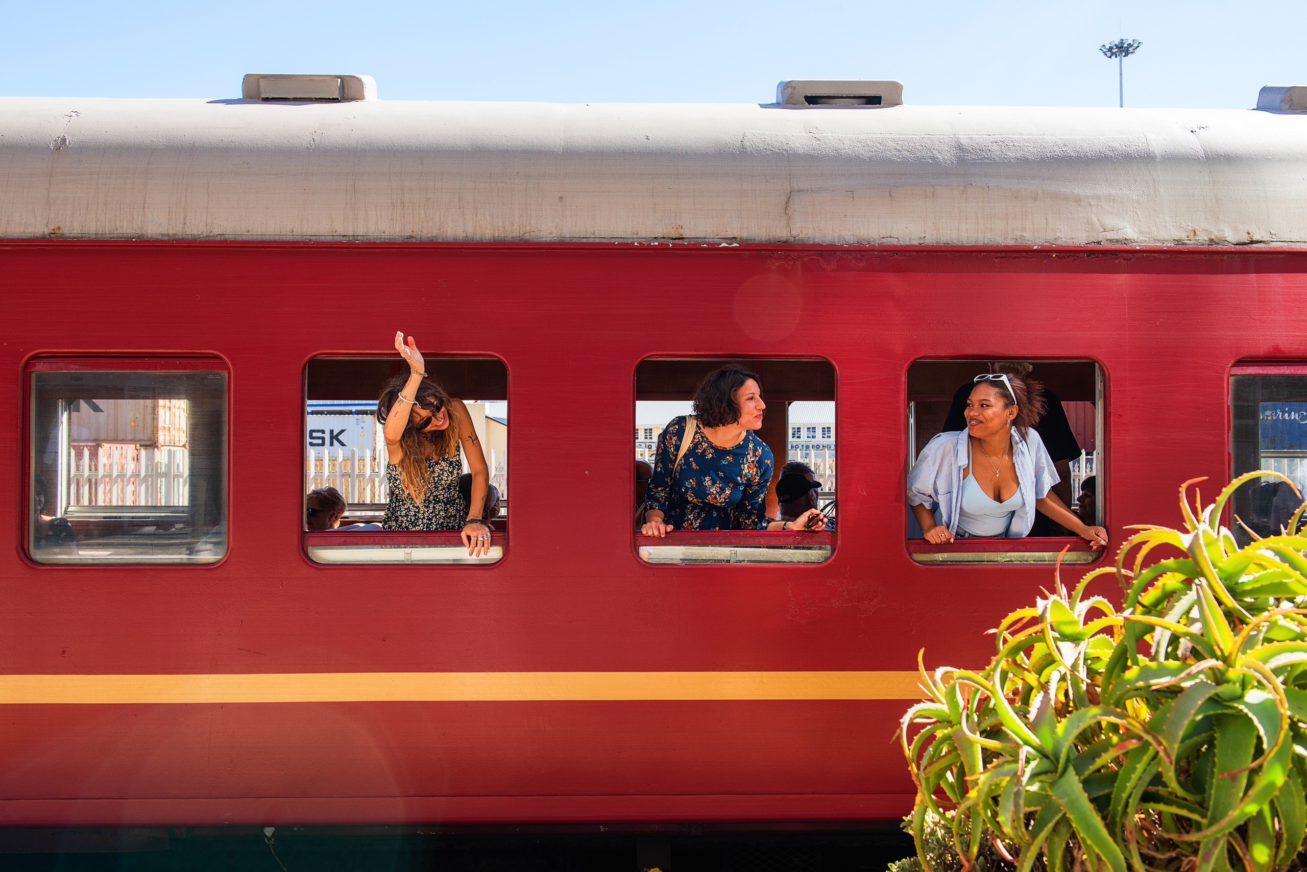 Three women wave and smile from windows of a red train car.