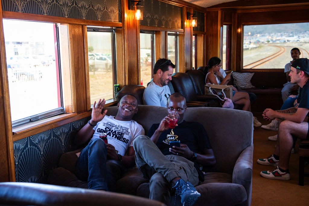 People sitting on a train, two men smiling and holding drinks in foreground.