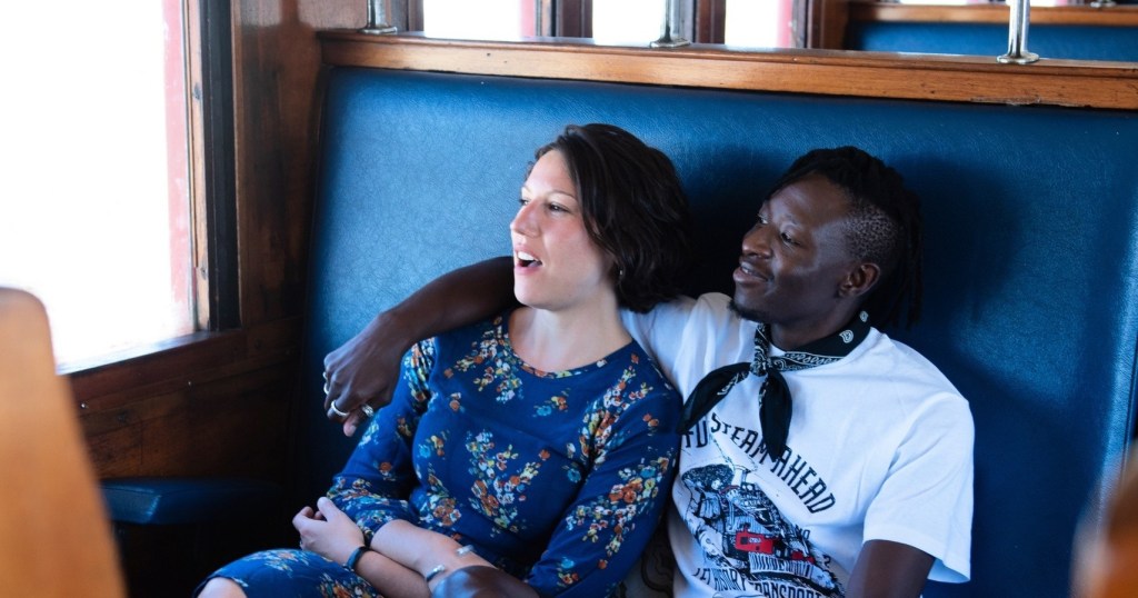 Two people sitting together on a train seat, one with an arm around the other.