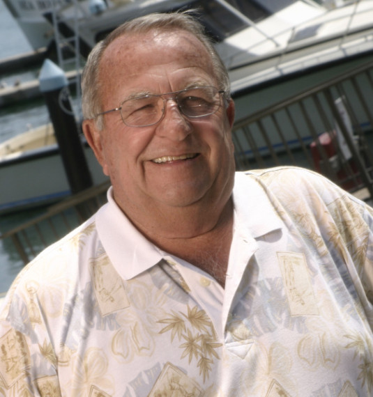 Smiling man in patterned shirt by marina with boats.