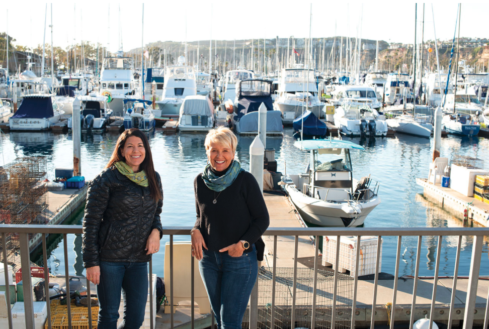 Two people smiling at a marina with boats docked in the background.