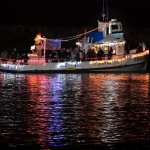 Boat decorated with festive lights reflecting on the water at night.